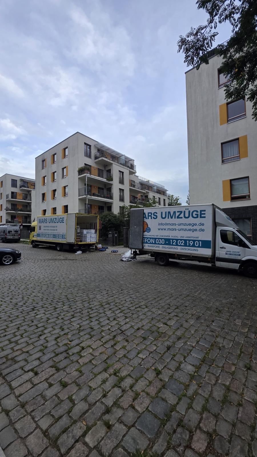 Ein Lkw mit geöffneter Ladefläche voller Umzugskartons und ein Transporter von Mars Umzüge stehen auf Kopfsteinpflaster vor einer Wohnanlage mit Balkonen