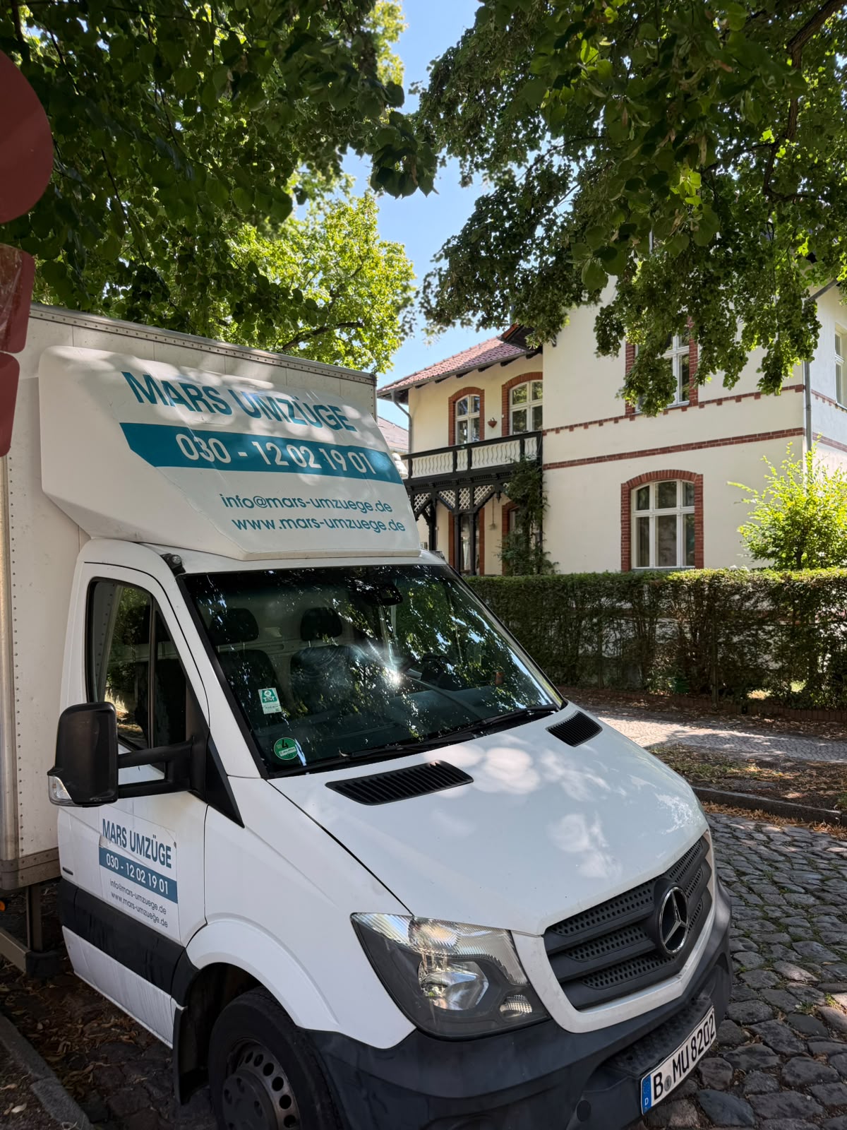 Umzugsfahrzeug von Mars Umzüge steht auf Kopfsteinpflaster vor einem hellen Altbau mit Backsteinfassung, Balkon und Hecke